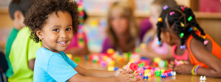 Image of young children building with colorful blocks in a classroom. Child in the foreground is smiling and looking at the camera.