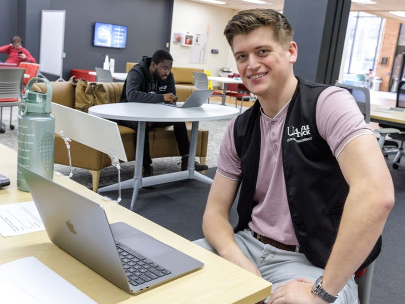 Male student tutor sitting at a desk with his computer in the campus Library.