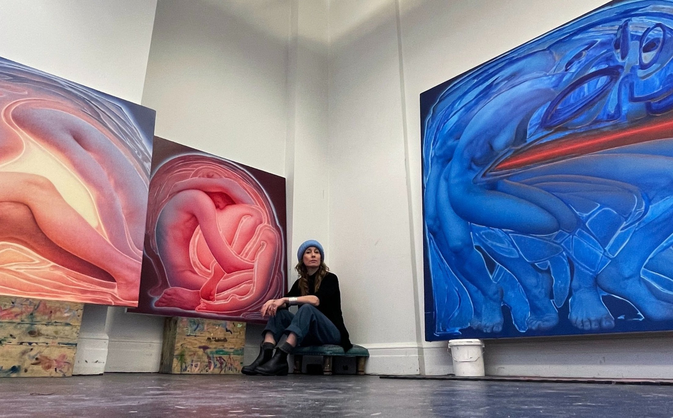 Artist Aleah Chapin sitting in a studio space with some of her paintings.