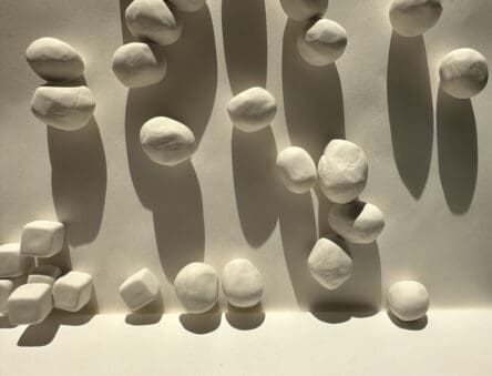 White, hand formed ovoids and square ceramic objects attached to a wall with overhead lighting casting long shadows beneath each object.