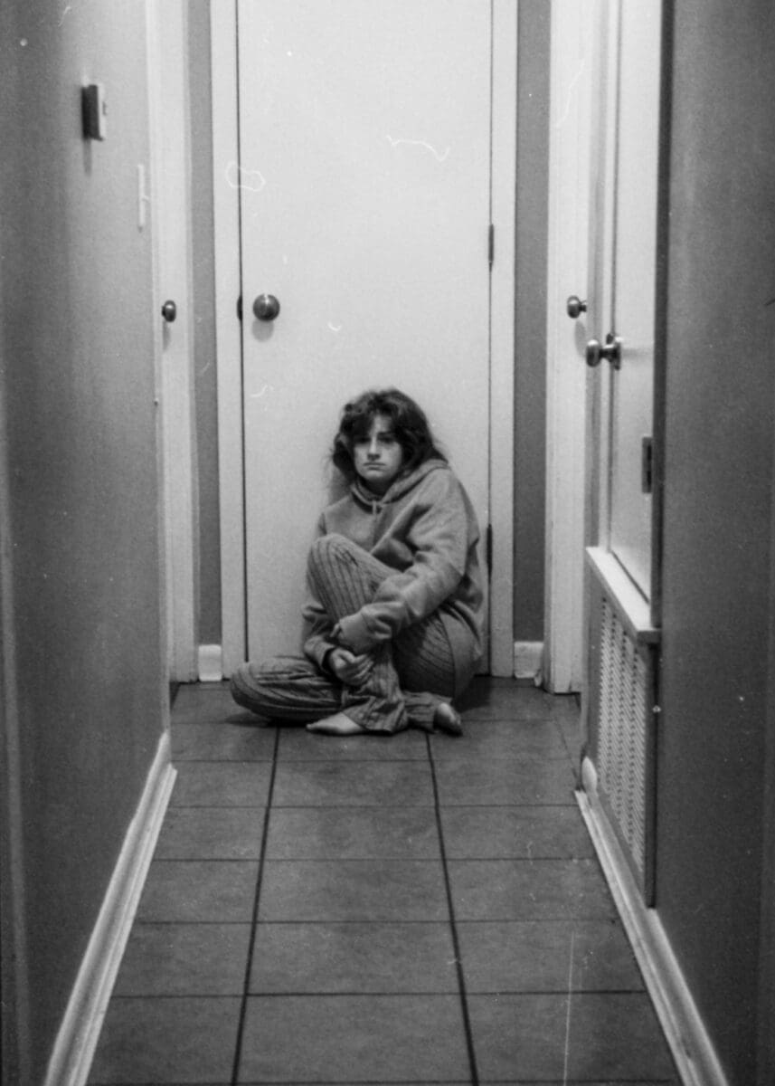 A self-portrait image of the artist sitting on the floor at the end of the hallway in her home.