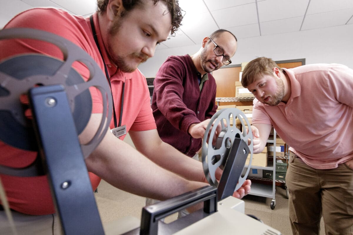 Three graduate assistants work on loading film into a 16mm film projector