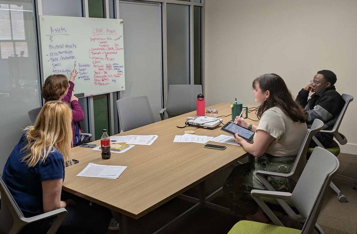 Three graduate students and an archivist sit at a conference table, looking at a white board. The archivist points to the white board which has two columns listing Assets and CAHC Assets