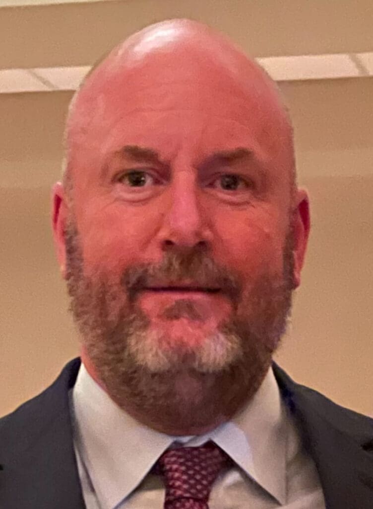A man with a beard wearing a blue suit and red tie smiles at the camera.