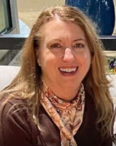 A woman with light hair wearing a white and red scarf and brown shirt smiles at the camera. 