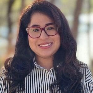 A woman with long brown hair, red glasses, and a striped shirt smiles at the camera.