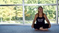 Ms. Cai Carvalhaes sitting cross legged on a blue yoga mat with a peaceful window and trees behind her.
