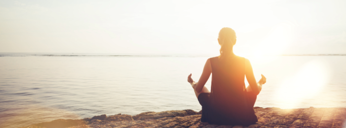 Person meditating by water as the sun rises