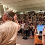 Bruce and a Scout leader stand in front of a large audience during the Merit Badge University opening ceremony as attendees raise the Scout sign.