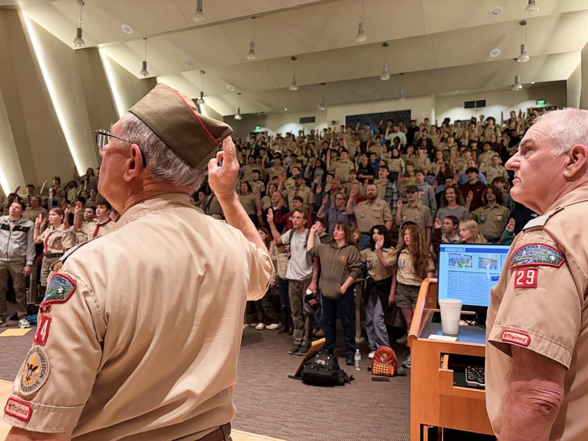 Bruce and a Scout leader stand in front of a large audience during the Merit Badge University opening ceremony as attendees raise the Scout sign.