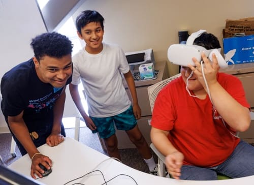 camp instructor leaned over using computer mouse smiling and giving guidance while student participant adjusts his virtual reality headset while a young participant watches and smiles
