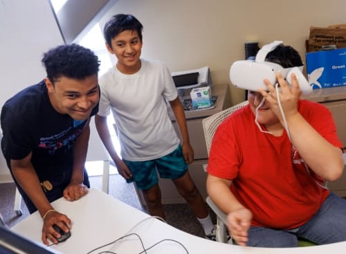 camp instructor leaned over using computer mouse smiling and giving guidance while student participant adjusts his virtual reality headset while a young participant watches and smiles