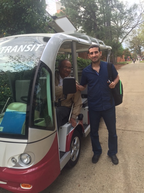 A male student stands on a sidewalk next to a small campus transit bus. The student and the bus driver are smiling.