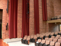 Photo of the Stella Boyle Concert Hall looking out from the stage to the auditorium. There is a tall brick wall with long hanging drapes on the side of the seating area where there are rows of cushioned seats, The hall is empty of people.