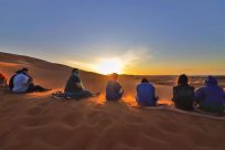 Photo of a group of 5 students sitting on sand dune in Morocco watching the sun set.
