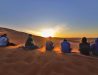 Photo of a group of 5 students sitting on sand dune in Morocco watching the sun set.