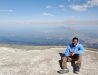 Photo of a male Study Abroad student sitting on a rock - behind is a panoramic view of mountains likely n a foreign country.