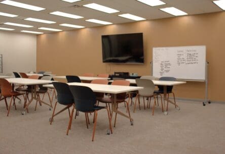 Image of room with desks, chairs, a white board and monitor.