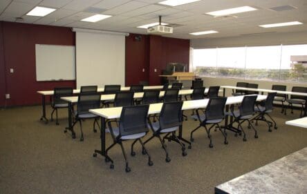 Image of a classroom with a screen, podium and tables and chairs.