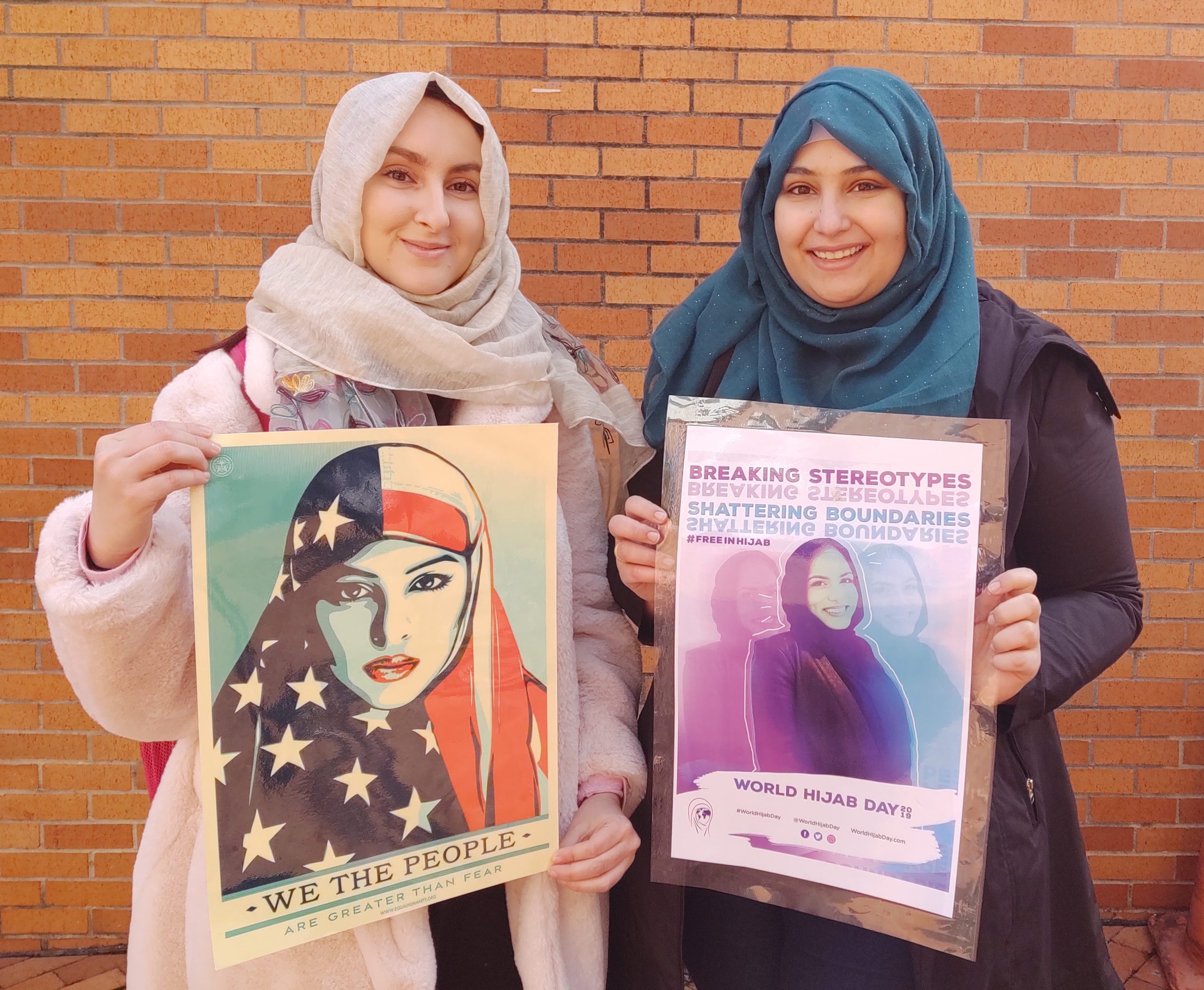 Two female students wearing the hijab traditional head covering hold World Hijab Day posters that read "We The People Are Greater Than Fear" and "Shattering Stereotypes, #FreeInHijab, World Hijab Day".