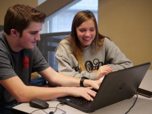 Pictured are two competitors from the hash code competition event sitting at a table looking at a computer together, smiling.