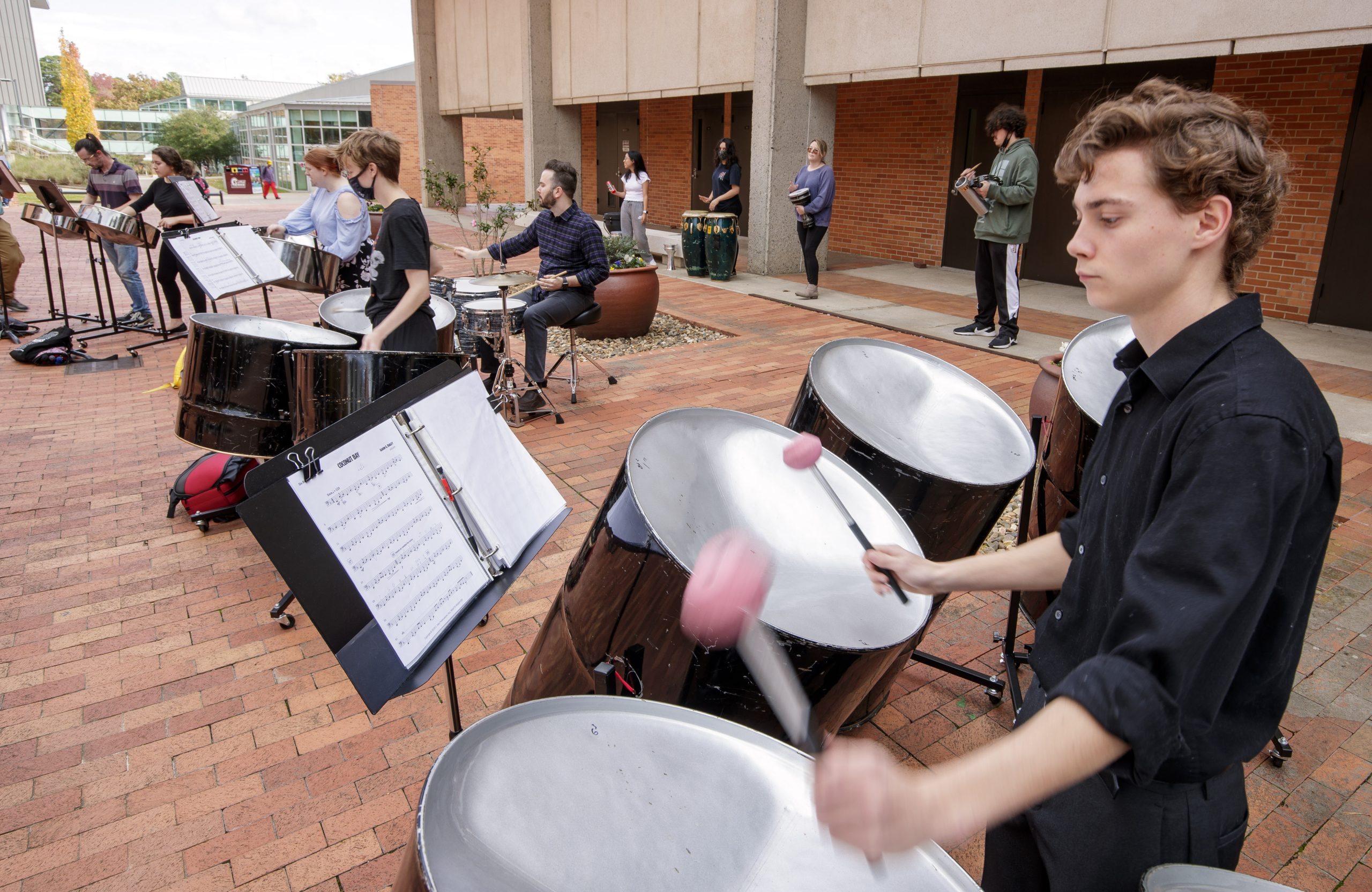 Six members of UA Little Rock's Trojan Steel Band playing instruments outdoors on campus.
