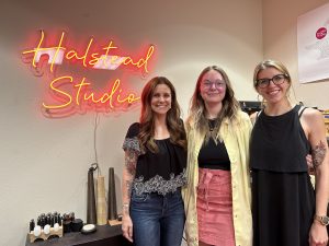 Three women pose in front of an LED sign that says "Halstead Studio."