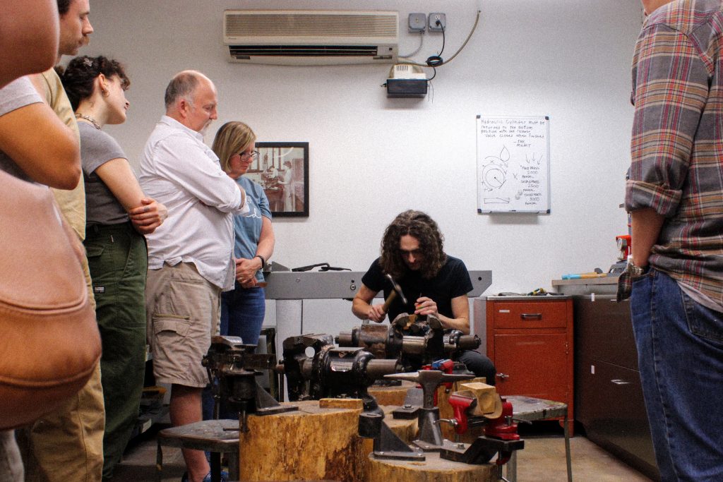 Four people stand on the left side of the image, while one person stands on the right. A teacher is holding a hammer and teaching a spoon-forging workshop.