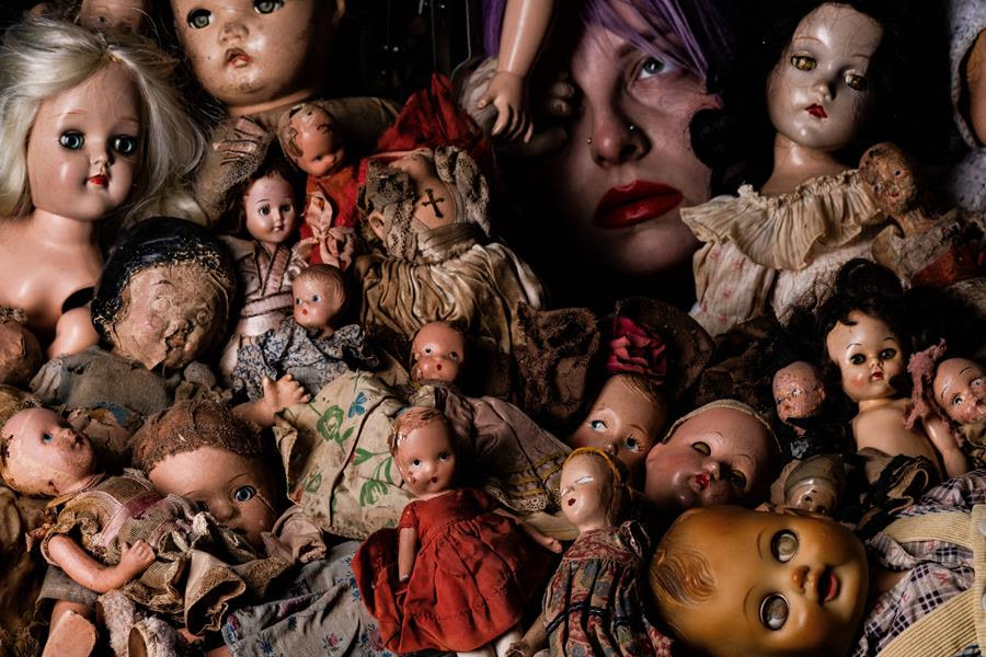 Photograph featuring 20+ broken dolls in various conditions with a woman's face poking out behind the dolls.
