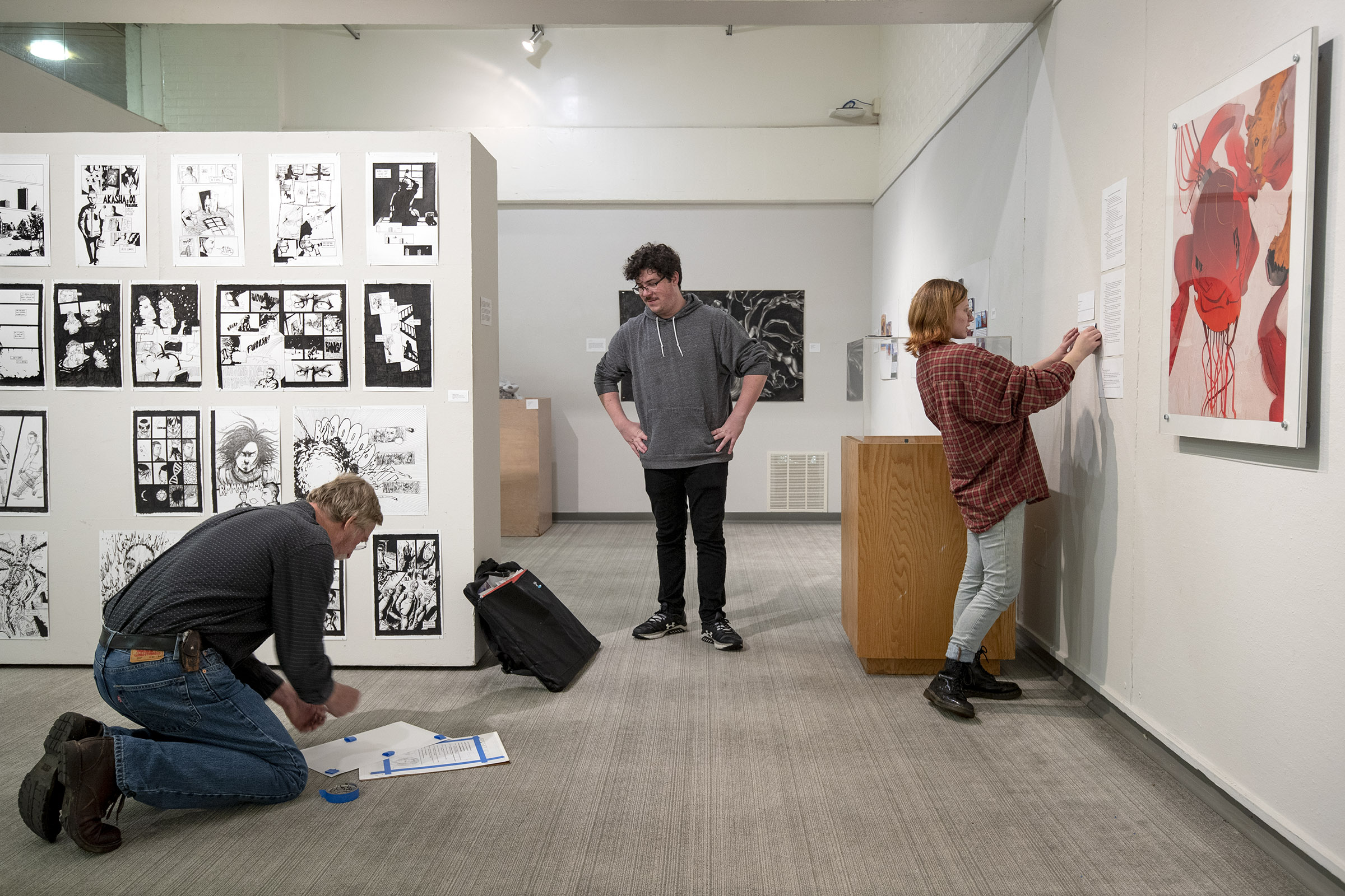 In an art gallery, on the left side, a man working on the ground tapes a piece of paper, while a student stands and watches. Another student on the right side tapes up a piece of paper on the gallery wall.