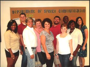 A group of ten students stand in front of a bulletin board that reads "Department of Speech Communication.: