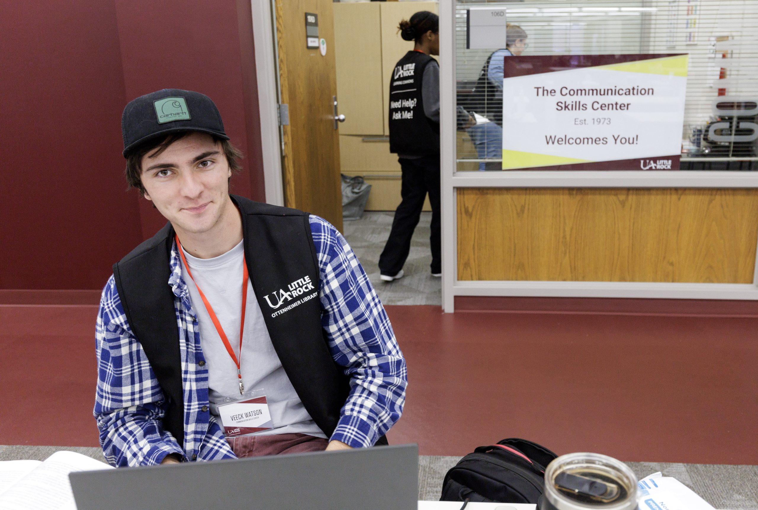 A student sits at a desk wearing a vest that notes he is a tutor. Behind him is a sign that reads "The Communication Skills Center, est. 1973."