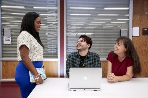 Two students sit at a desk while another stands off to the side. The three are having a conversation in the Ottenheimer Library at UA Little Rock.