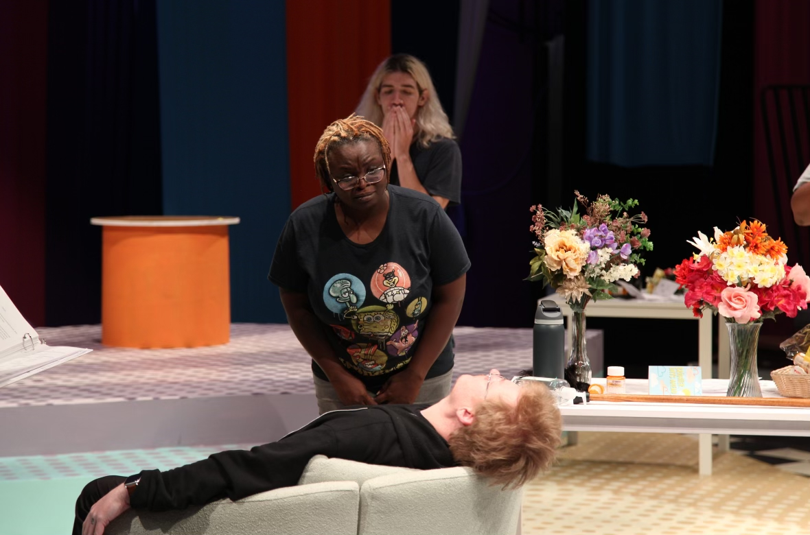 On a stage, an actor stands in the background with their hand over their mouth. Another actor leans over someone who is lying out in a chair.