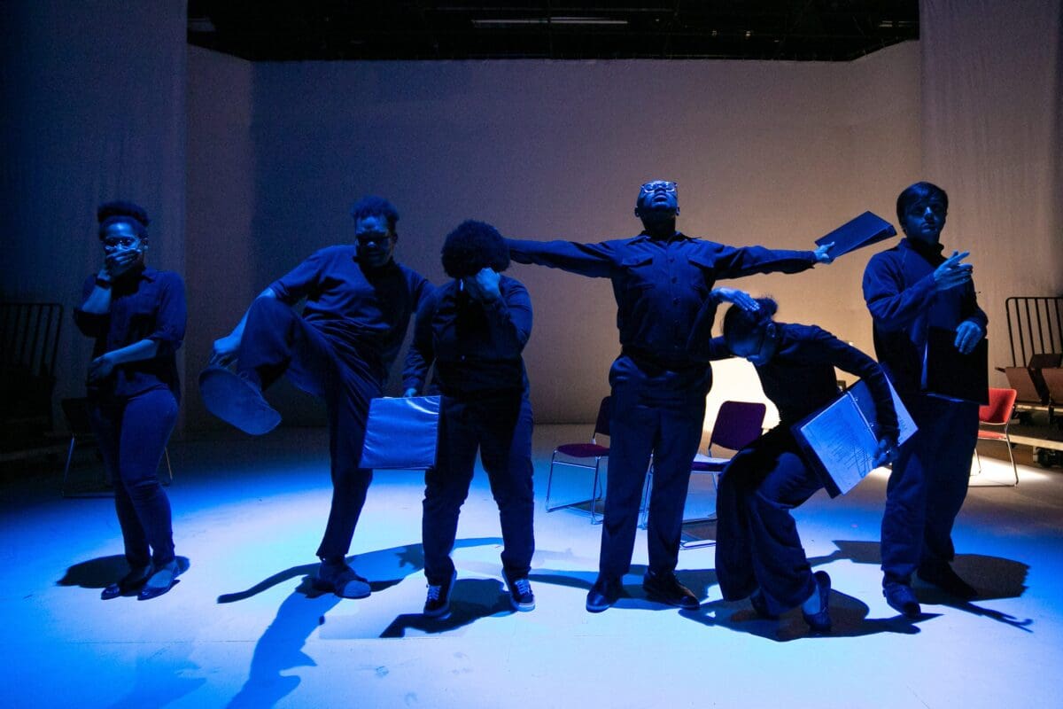 Six students stand in a row on stage each in different poses, holding binders with their scripts.