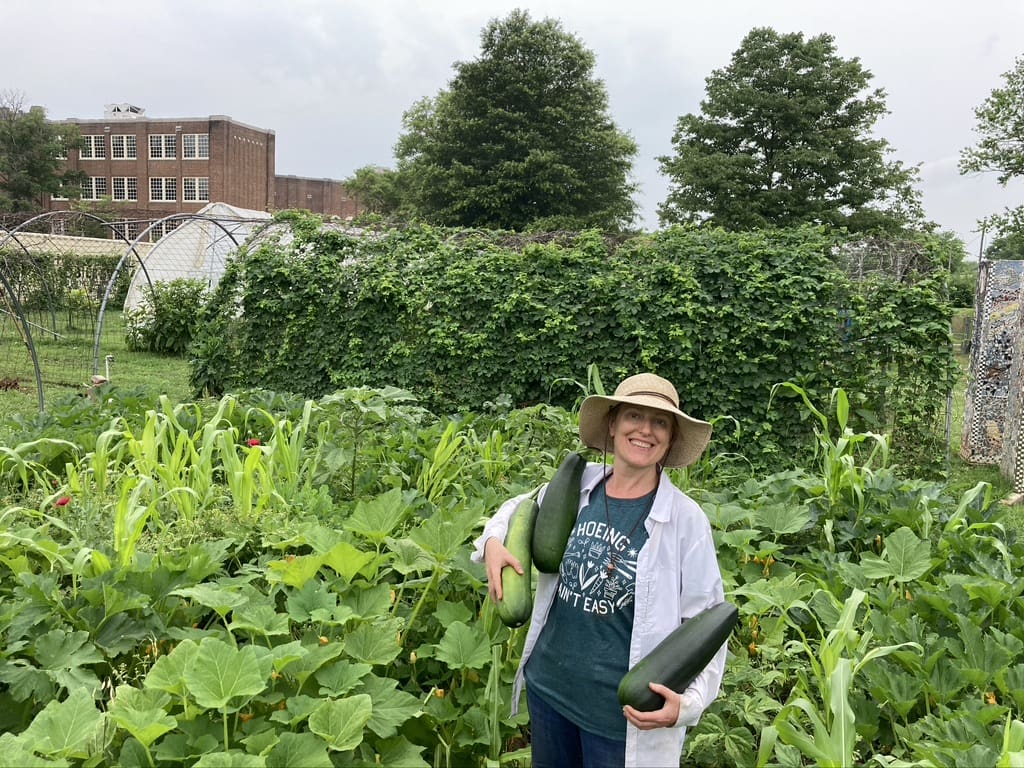 A woman poses with three large zucchini in the middle of a garden.