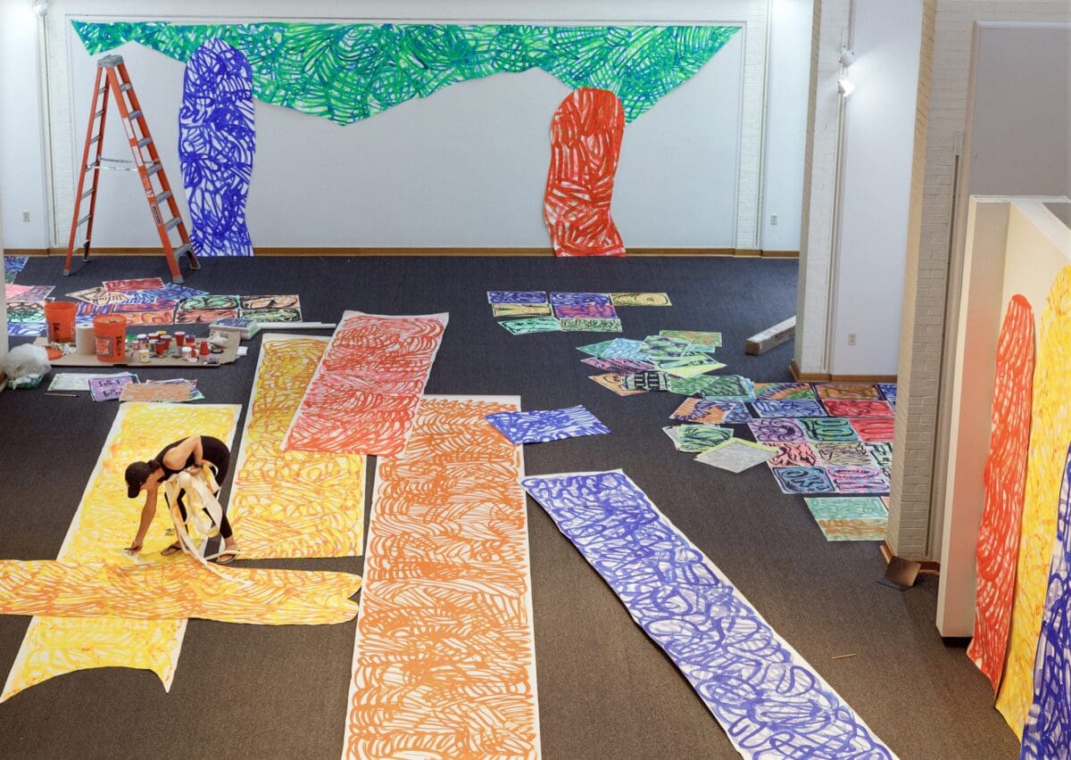 An aerial shot, a woman stands over a long piece of paper in a gallery with scissors in her hands. Scraps of paper are in another hand, and she's standing around longs trips of colorful paper.