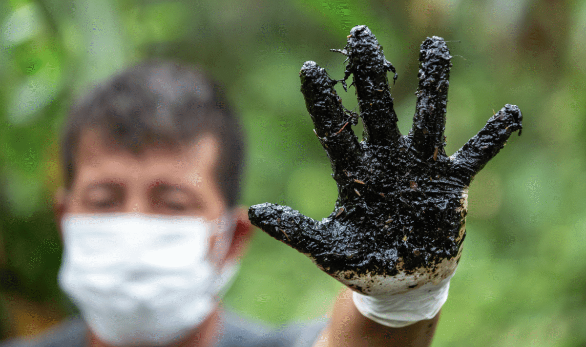A masked person wearing a glove raises a hand coated in black oil residue, illustrating environmental contamination.