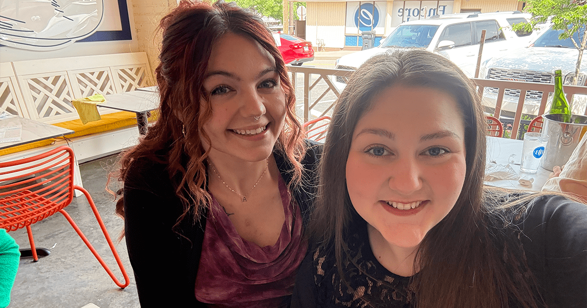 Two women smile for a selfie while seated at a restaurant table. One person has curly red hair and wears a purple top with a black cardigan, while the other has long dark hair and wears a black lace-accented top. The bright, casual dining space features colorful chairs, large windows, and a view of parked cars outside.