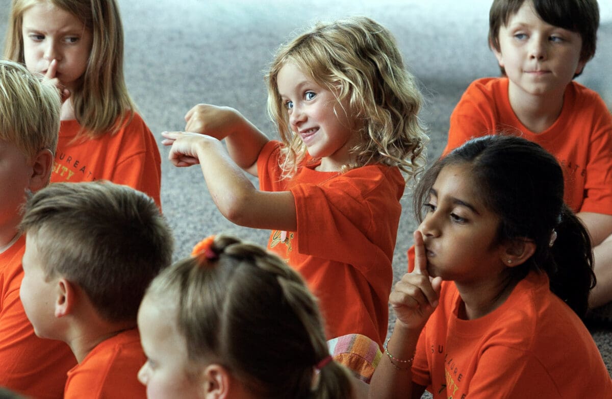 A diverse group of elementary children sit in a circle wearing orange T-shirts, while participating in the Summer Laureate program at UA Little Rock.